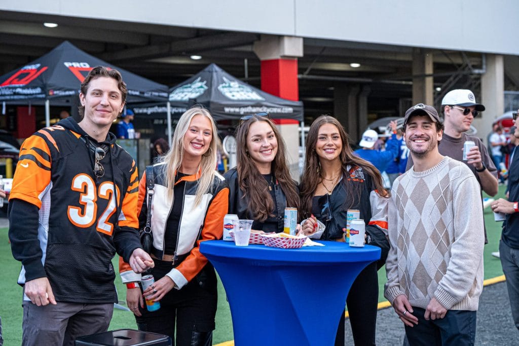 Five people, some in sports jerseys, smile around a blue table with drinks at an outdoor tailgate party NYC. Tents and other partygoers can be seen in the background, capturing the city’s vibrant tailgating spirit.